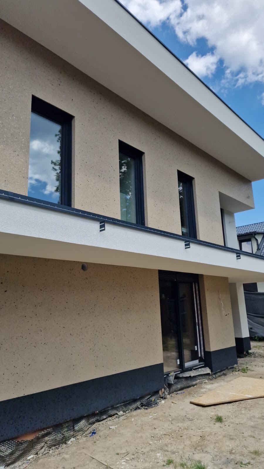 Elewacja domu jednorodzinnego z oknami w czarnych ramach i wykończeniem w odcieniach beżu i bieli. Widoczne fragmenty nieba i sąsiedniego budynku.