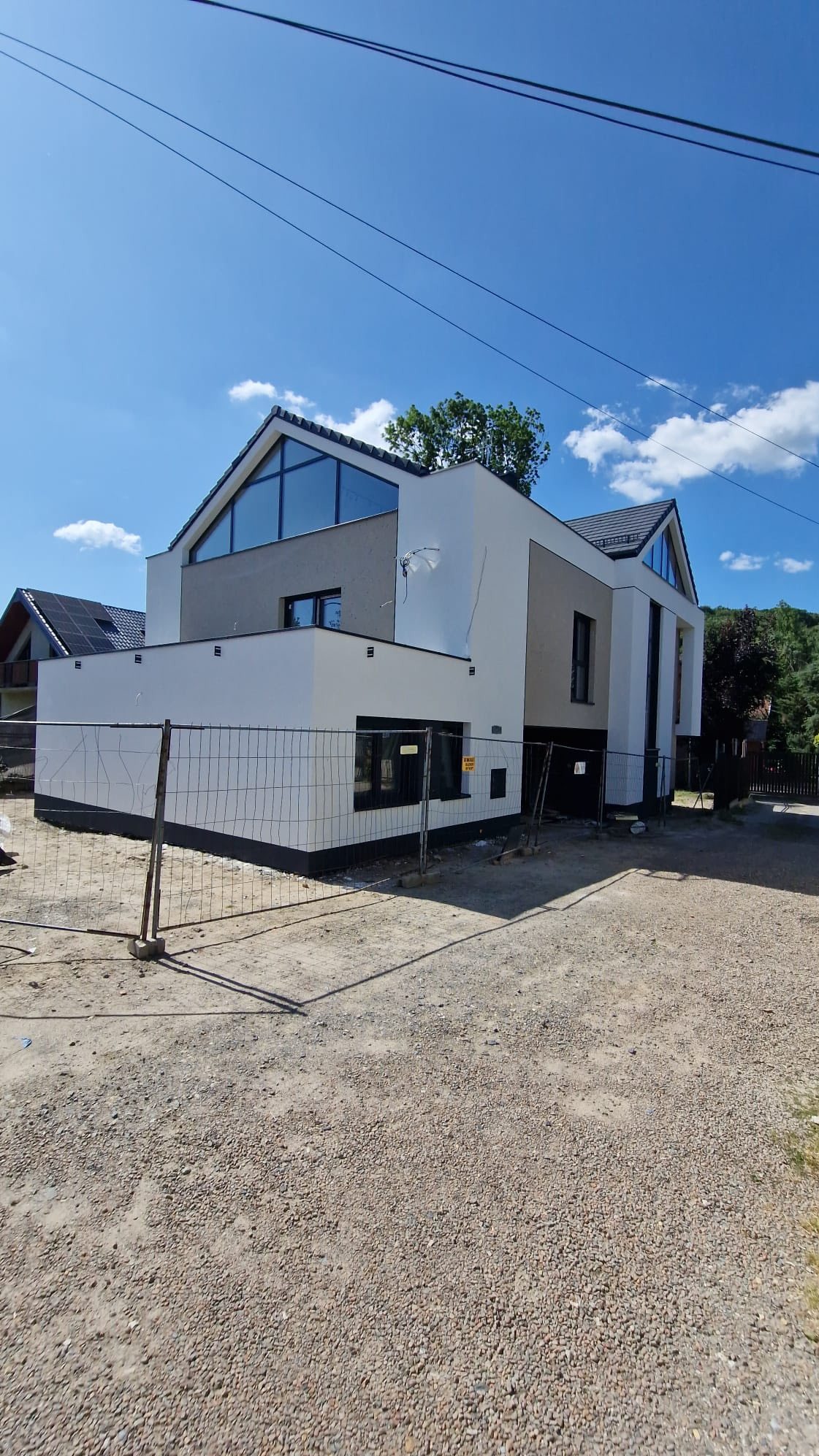 Nowoczesny dom jednorodzinny z dużymi oknami, wykończony w bieli i szarości, ogrodzony siatką. Widoczne elementy elewacji i otoczenie w słoneczny dzień.