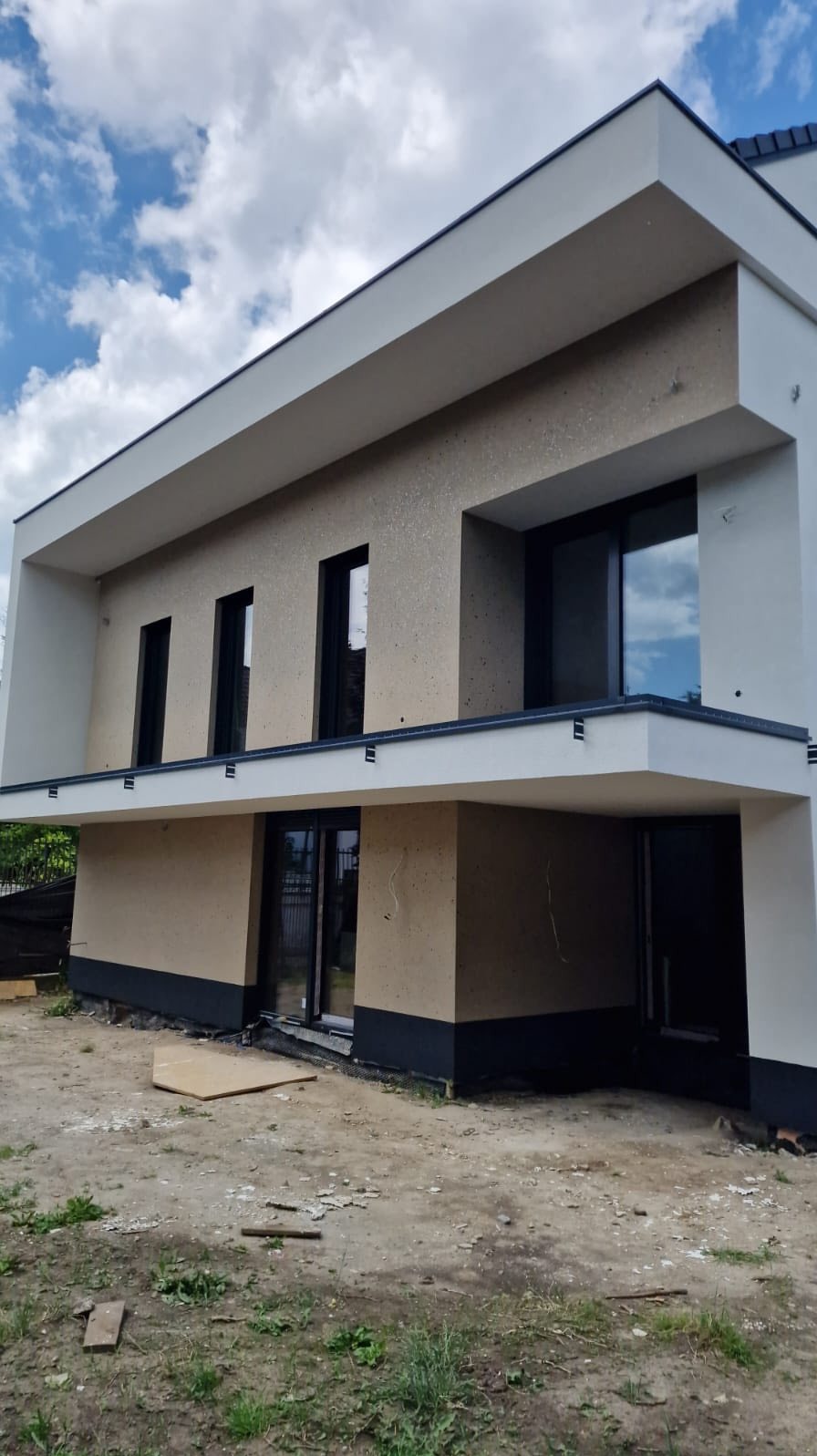 Dwukondygnacyjny dom z jasną elewacją i ciemnymi ramami okiennymi, widziany pod kątem. Tynk strukturalny na ścianach, widoczne elementy wykończeniowe.