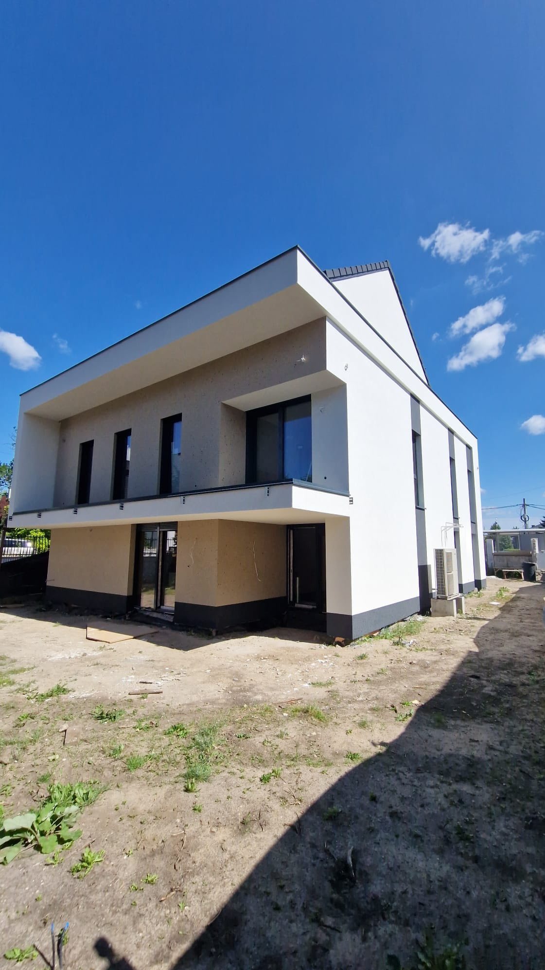 Nowoczesny, dwukondygnacyjny dom z białymi ścianami i ciemnymi oknami, częściowo wykończony, z widocznym ociepleniem na parterze i błękitnym niebem w tle.