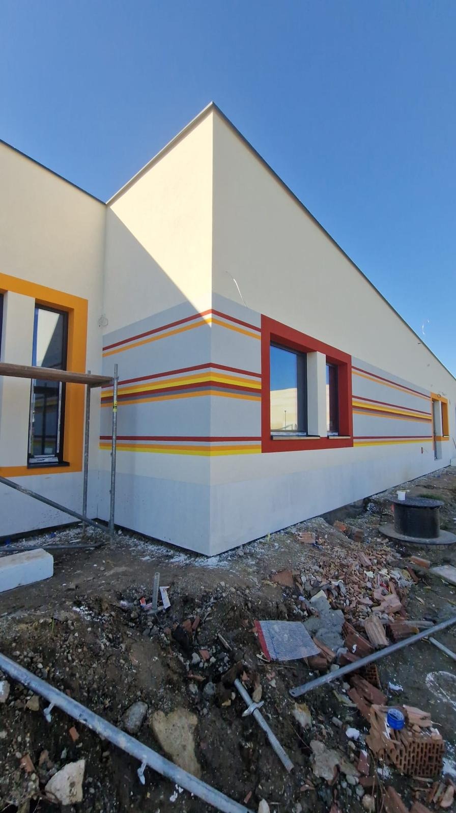 Elewacja budynku po malowaniu z dekoracyjnymi, kolorowymi pasami i obramowaniami okien. Widoczne rusztowanie i teren budowy.