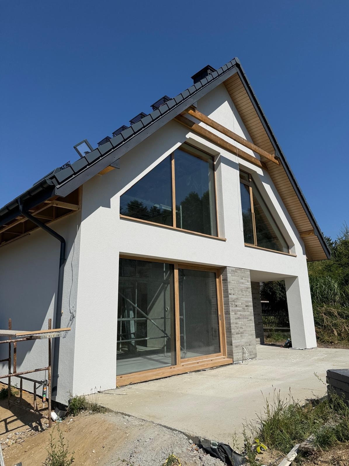 Nowoczesny dom jednorodzinny z białym tynkiem i dużymi oknami o nietypowym kształcie. Widoczne rusztowania sugerują prace wykończeniowe.