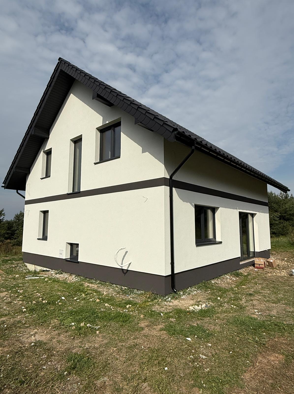 Dwukondygnacyjny dom z jasną elewacją i ciemnymi akcentami (okna, dach, pasy). Widoczne rury spustowe i fragment terenu przed budynkiem. Ujęcie z dołu, podkreślające bryłę.