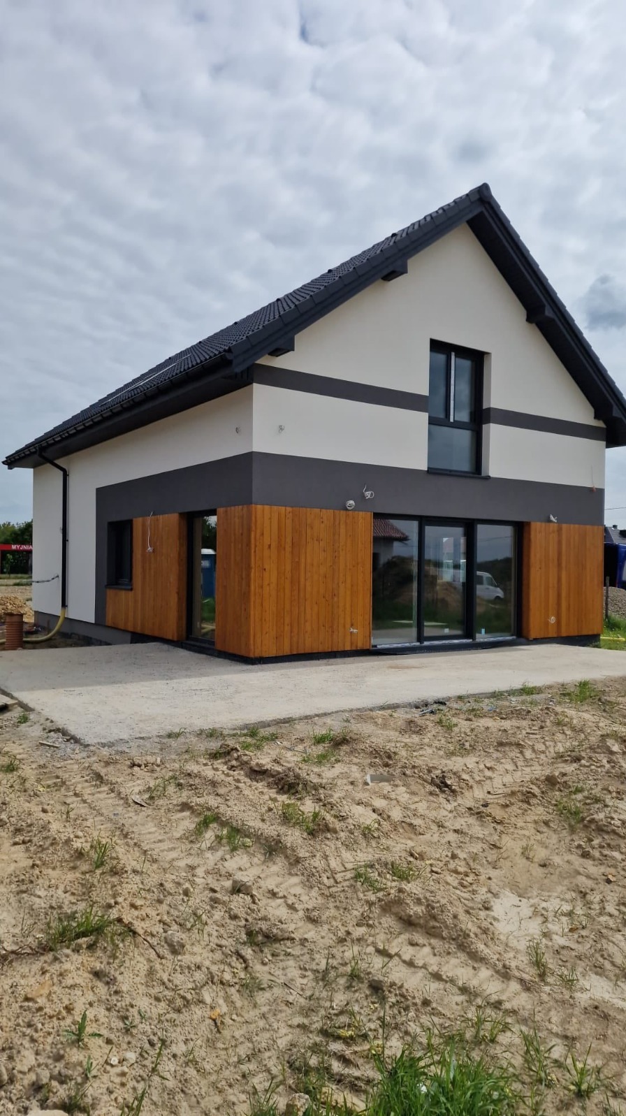 Nowoczesny dom jednorodzinny z drewnianą elewacją i dużymi oknami, wykończony w odcieniach bieli, szarości i brązu, na tle pochmurnego nieba.