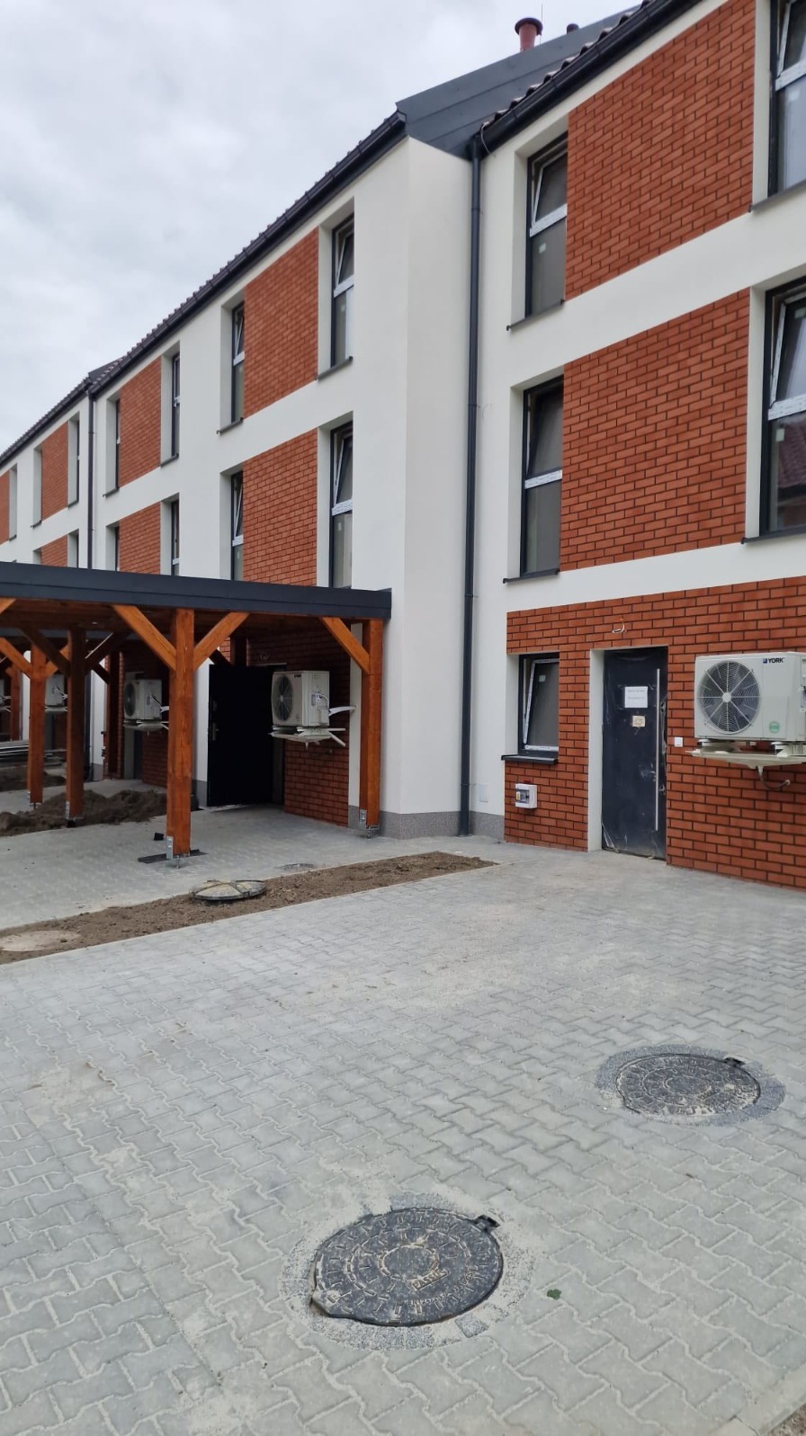 Nowoczesny budynek z elewacją z cegły i białymi elementami, widoczne klimatyzatory na zewnątrz, kostka brukowa na podwórzu i drewniana wiata.