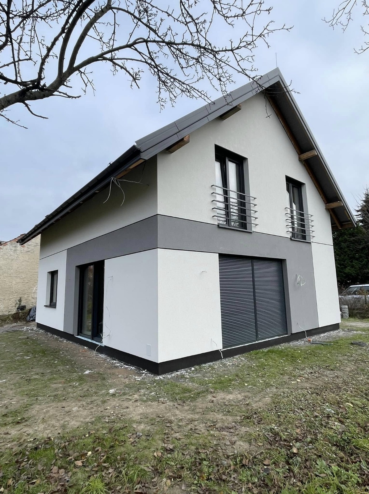 Dwukondygnacyjny dom z szarą, dwukolorową elewacją i czarnymi ramami okiennymi, widziany z perspektywy narożnej. Widoczne balustrady balkonowe i rolety zewnętrzne.