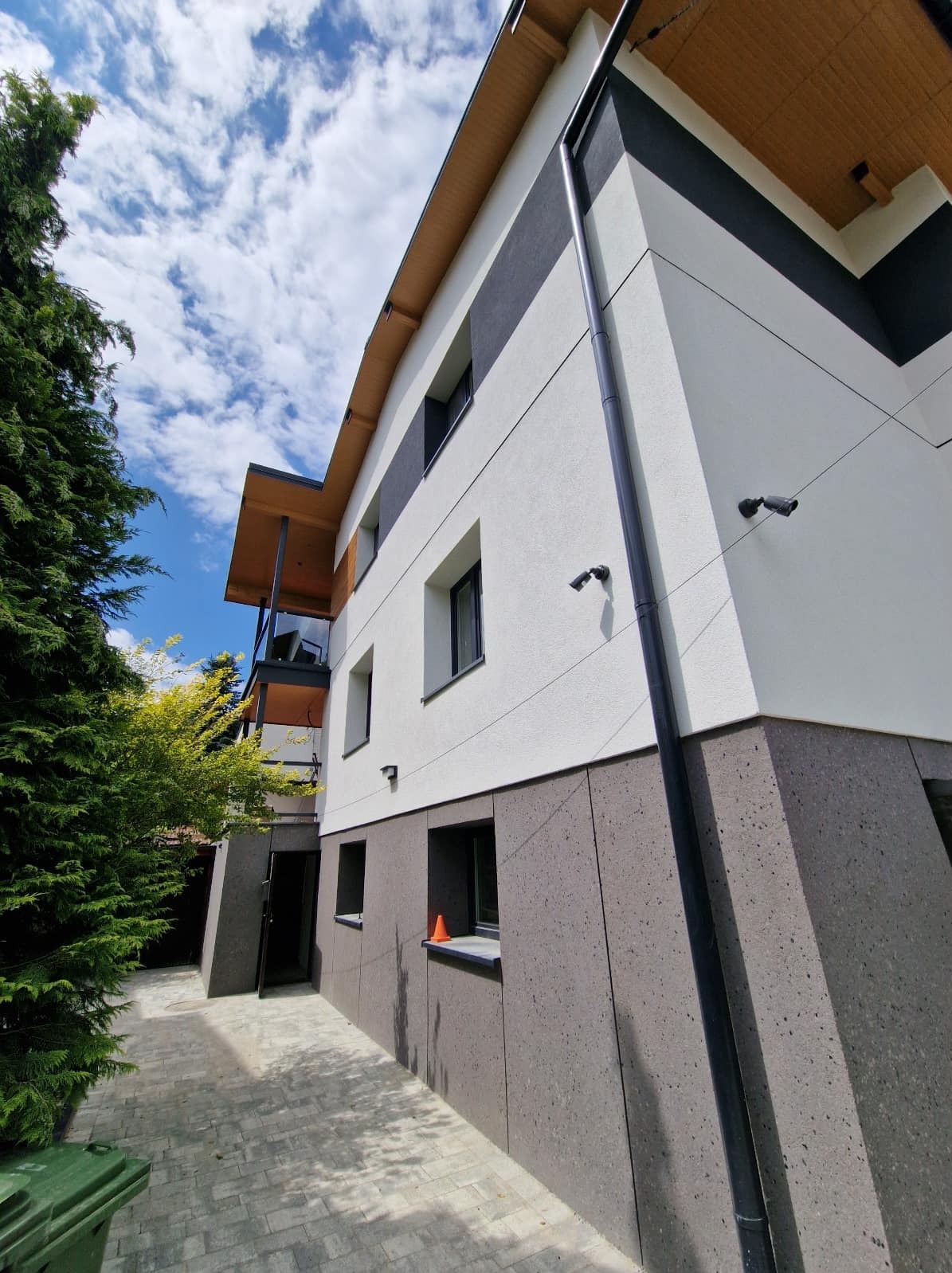 Nowoczesny budynek z białą elewacją i szarym cokołem, widok z dołu, błękitne niebo z chmurami w tle. Widoczne kamery monitoringu i elementy wykończenia.