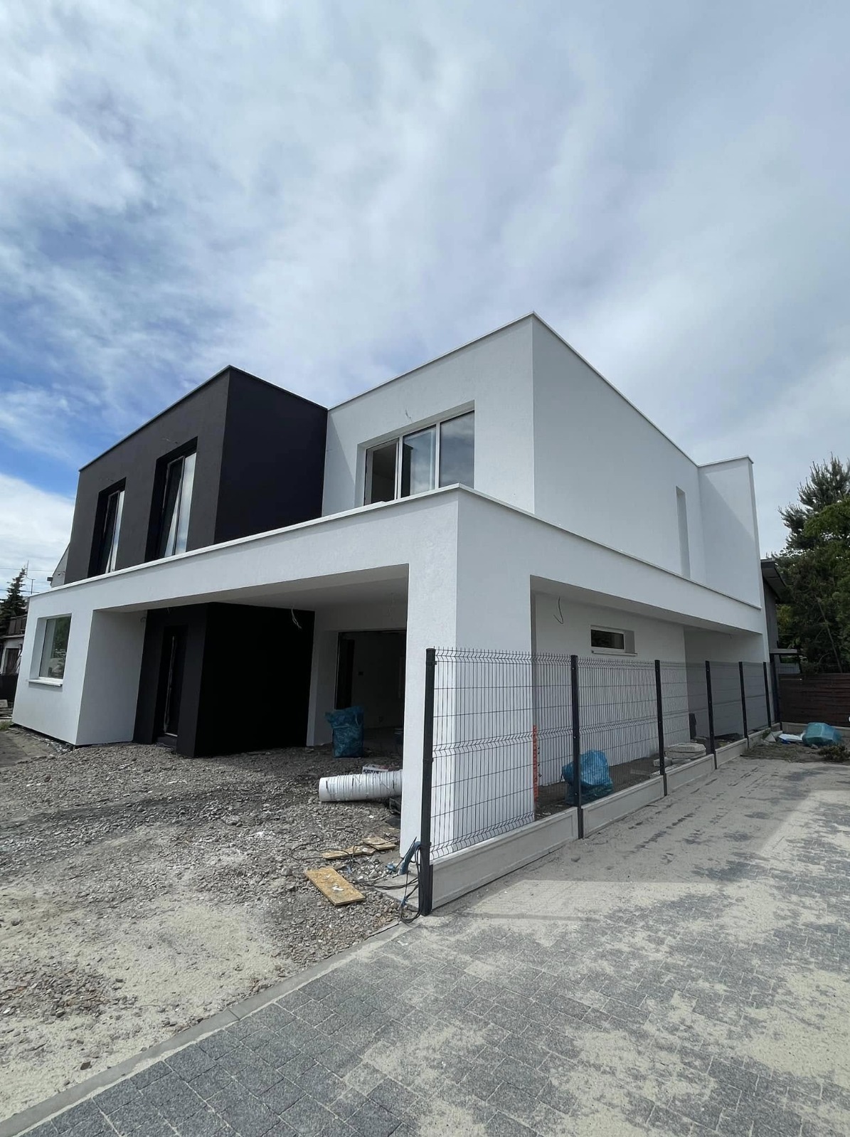 Nowoczesny, dwukondygnacyjny dom z czarno-białą elewacją, widoczny z zewnątrz. Częściowo wykończony teren wokół budynku. Metalowe ogrodzenie.