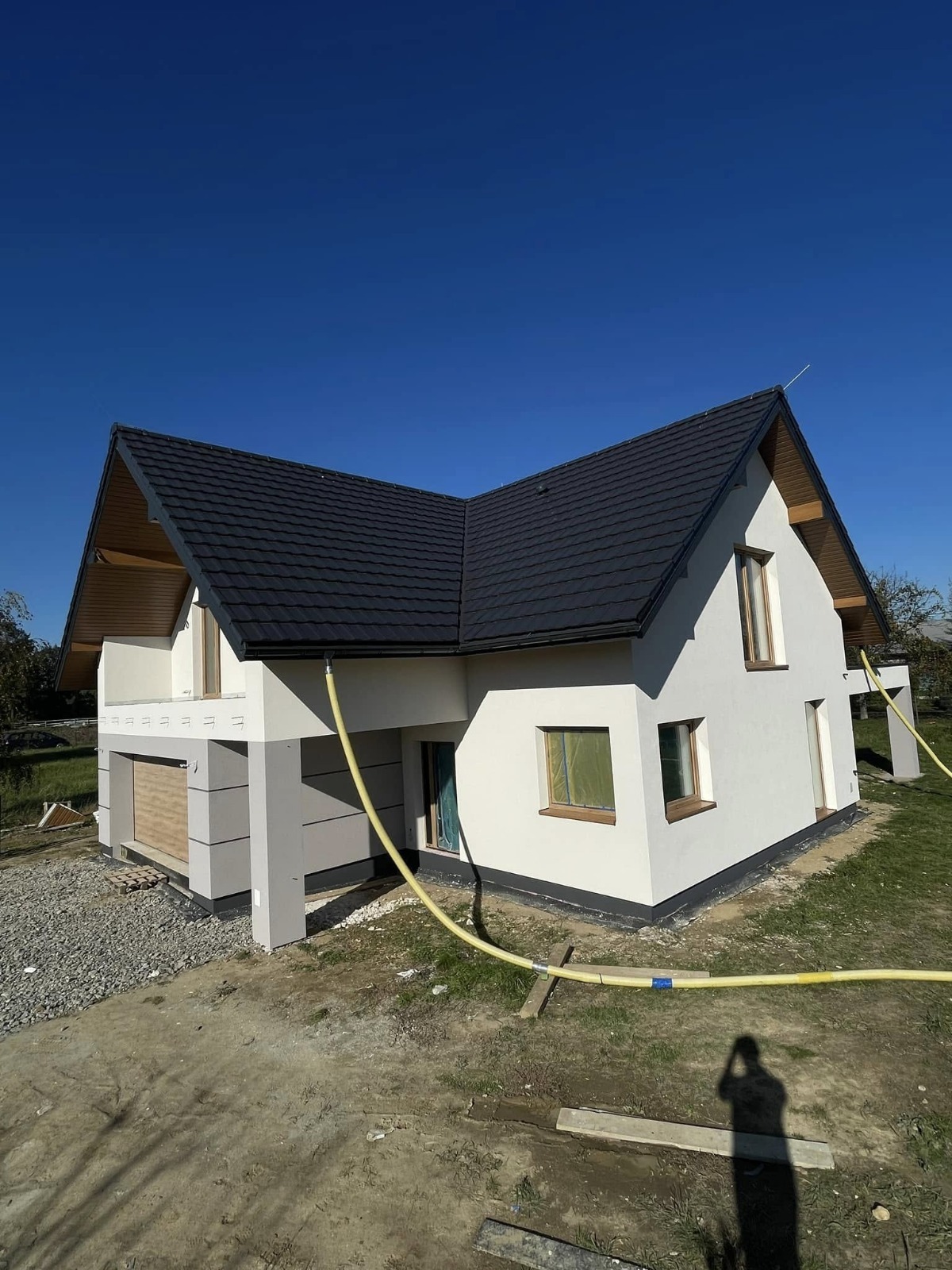 Nowoczesny dom jednorodzinny z ciemną dachówką i jasną elewacją, częściowo wykończony, z widocznymi wężami do prac elewacyjnych. Błękitne niebo w tle.