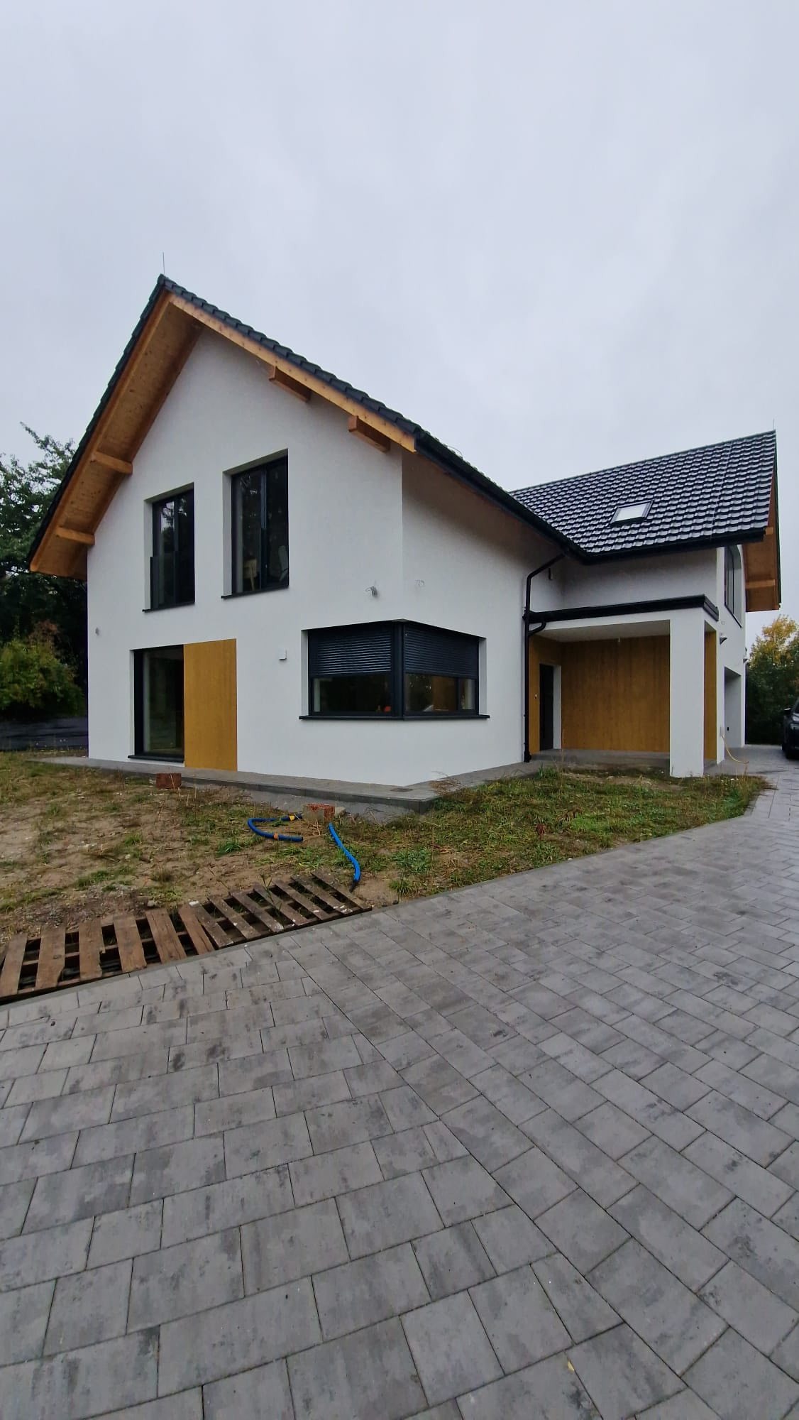 Nowoczesny dom z białą elewacją, czarną stolarką okienną i drewnianymi akcentami. Widoczny fragment brukowanego podjazdu i trawnika. Pochmurne niebo w tle.