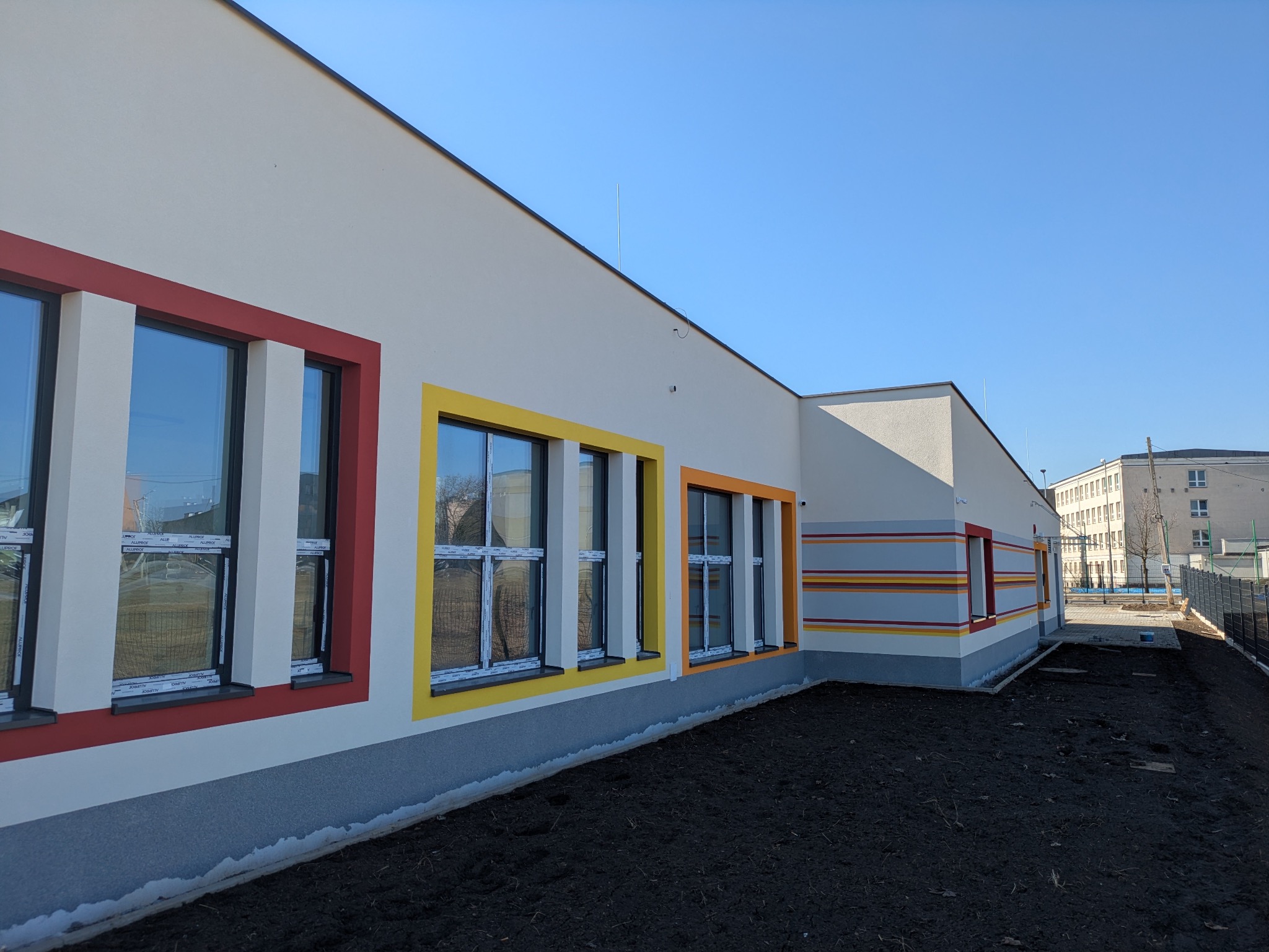 Nowoczesna elewacja budynku z kolorowymi obramowaniami okien: czerwonym, żółtym i pomarańczowym. Szara podmurówka i pasy dekoracyjne. Błękitne niebo w tle.