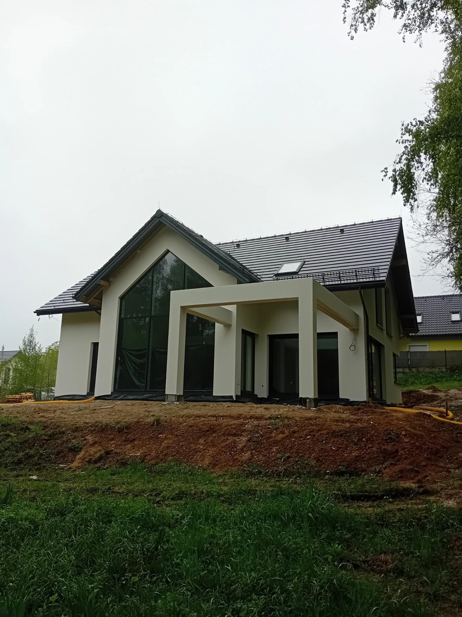 Nowoczesny dom jednorodzinny z białymi ścianami i dużymi oknami, częściowo zakryty folią ochronną, na etapie wykończenia elewacji. Widoczne elementy konstrukcyjne.