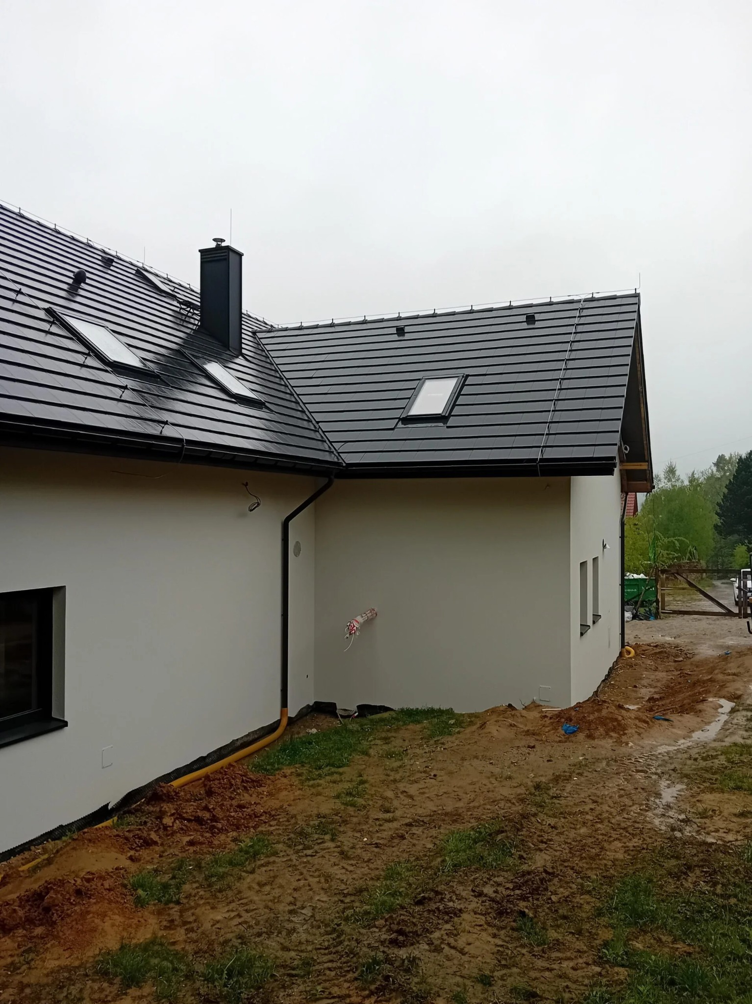 Biała elewacja domu jednorodzinnego z czarną dachówką i oknami dachowymi, widok z boku na tle pochmurnego nieba i błotnistego terenu.