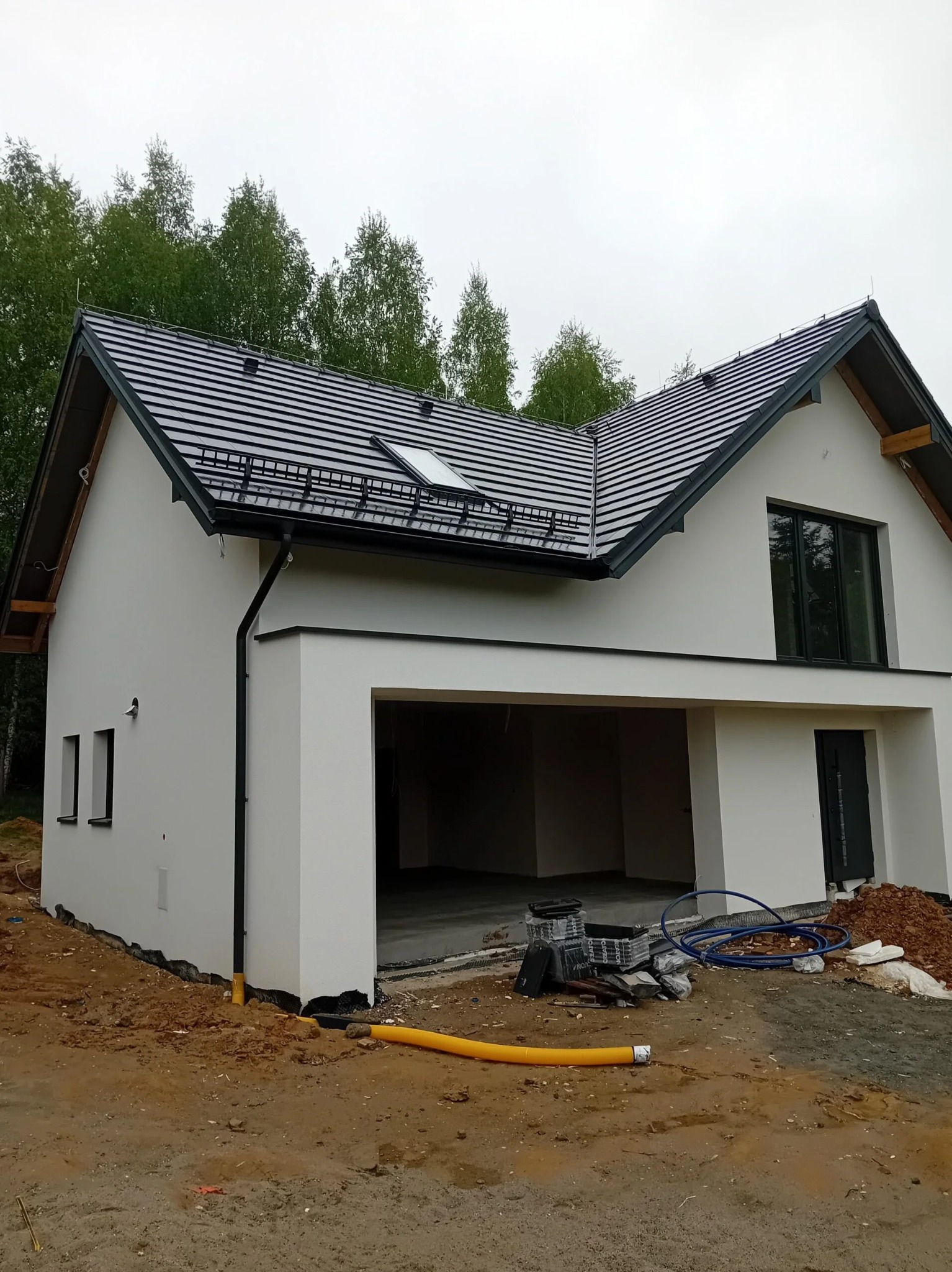 Biały dom jednorodzinny z garażem w trakcie wykończenia elewacji, widoczne rury i materiały budowlane na zewnątrz. Dach pokryty ciemną dachówką.