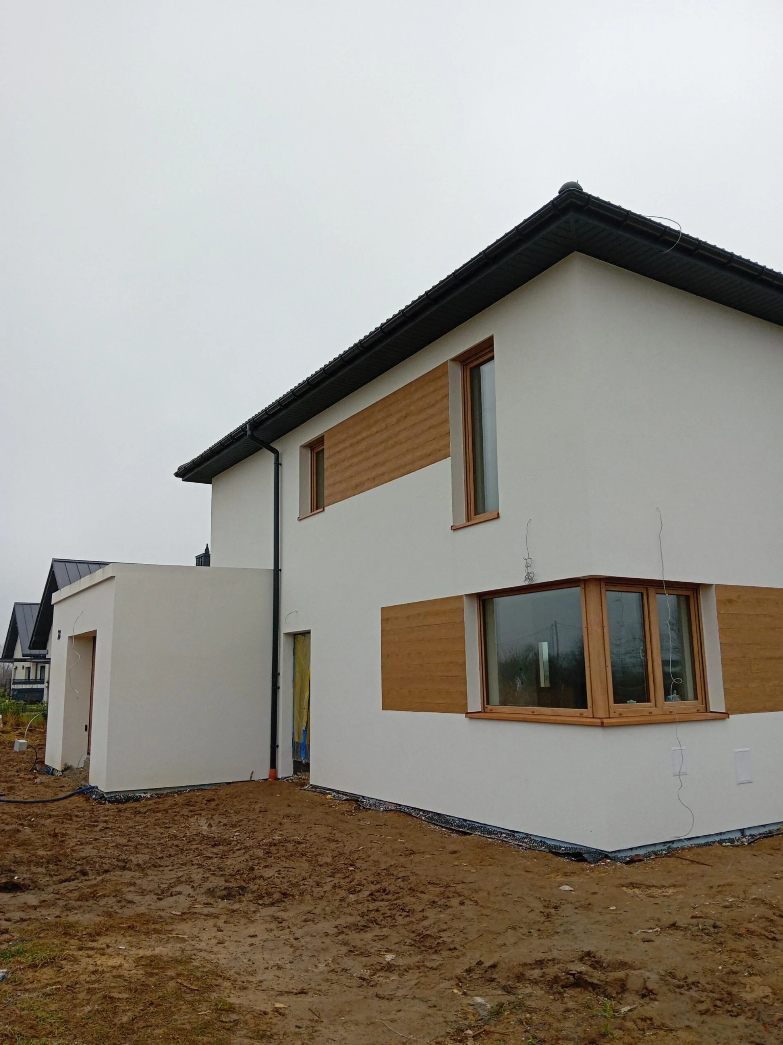 Nowoczesny dom z białą elewacją i drewnianymi akcentami wokół okien. Widoczne detale wykończenia i otoczenie domu w trakcie prac ziemnych.