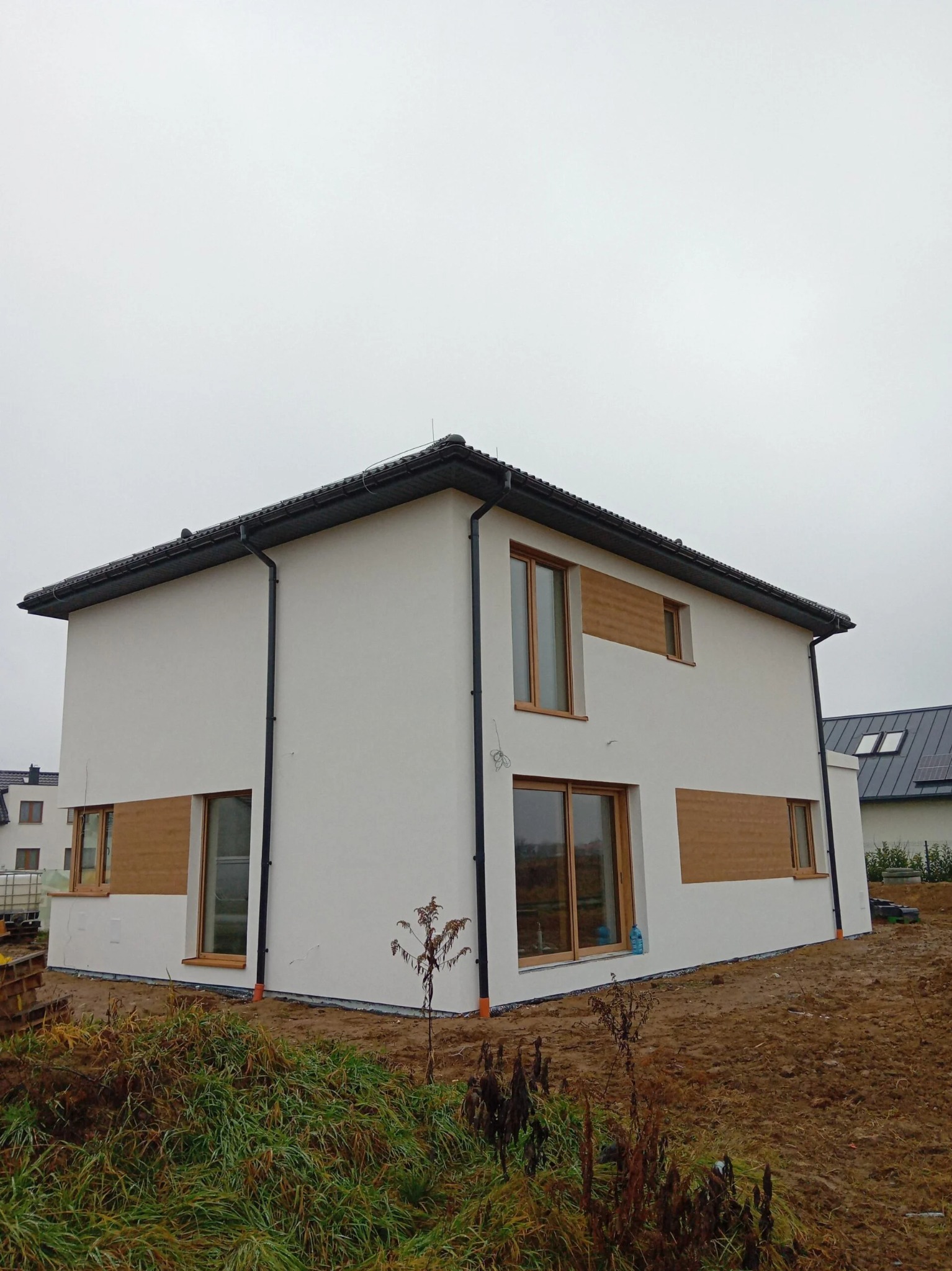 Dwukondygnacyjny dom z białą elewacją i drewnianymi akcentami, widoczny z zewnątrz na tle pochmurnego nieba i przygotowanego terenu.