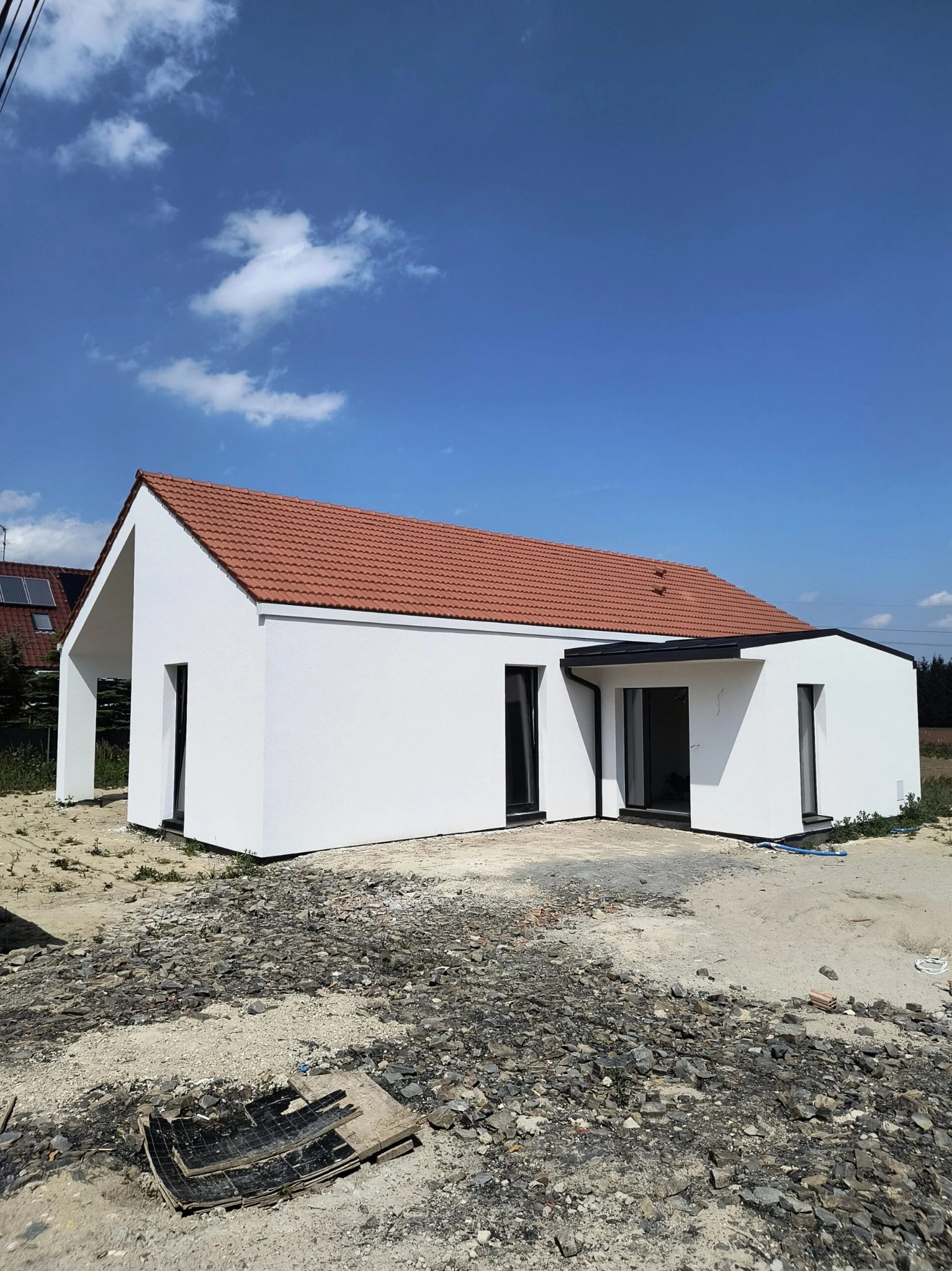 Biały dom jednorodzinny z czerwoną dachówką, widoczny w słoneczny dzień. Minimalistyczna bryła, czarne ramy okienne, surowe otoczenie placu budowy.