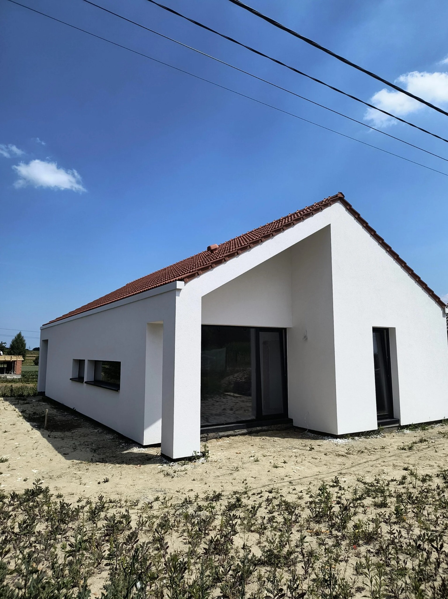 Biały, parterowy dom z czerwonym dachem w nowoczesnym stylu. Minimalistyczna bryła, duże okna, jasne tło nieba. Widok z poziomu gruntu, zieleń w pierwszym planie.