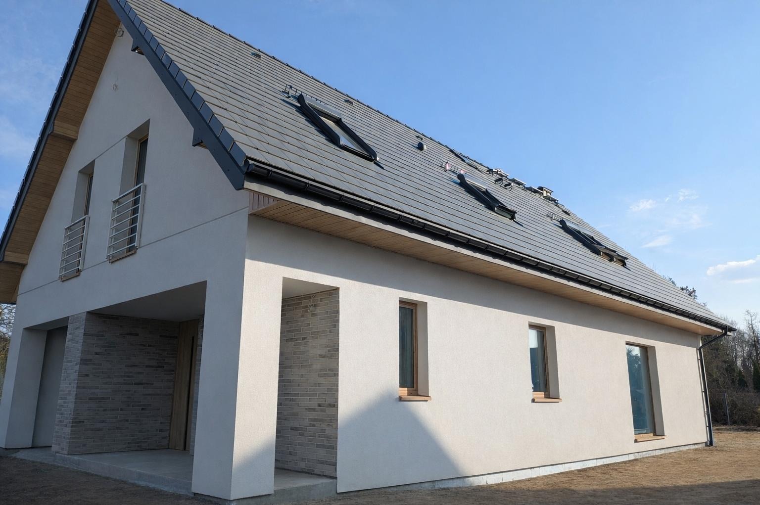 Nowoczesny dom jednorodzinny z jasną elewacją, grafitowym dachem z oknami połaciowymi i elementami z cegły klinkierowej. Widok z zewnątrz w słoneczny dzień.
