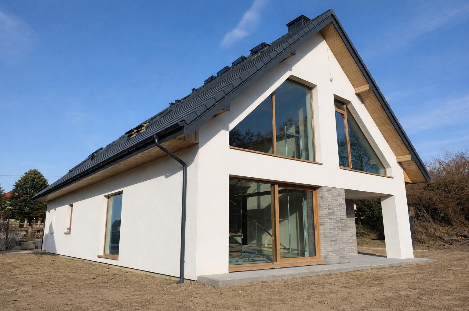 Nowoczesny dom jednorodzinny z białą elewacją i dużymi oknami o drewnianych ramach, widok z zewnątrz w słoneczny dzień. Część dachu jeszcze nieukończona.
