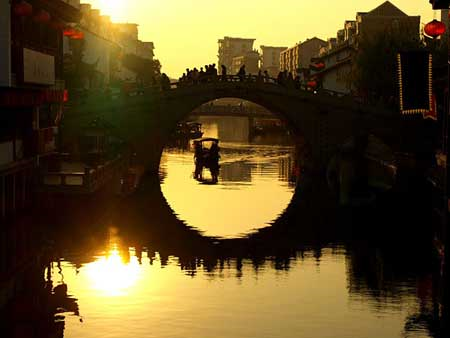 Złocisty krajobraz wodny z łodzią przepływającą pod kamiennym mostem w Chinach, oświetlony zachodzącym słońcem.