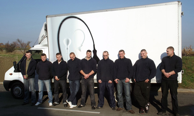 Zespół ośmiu mężczyzn w ciemnych bluzach i jeansach stoi przed białym busem z grafiką przypominającą słuchawkę telefoniczną na boku.