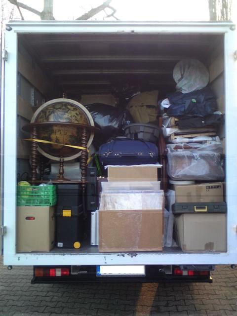 Wnętrze białego busa wypełnione po brzegi różnorodnymi przedmiotami podczas przeprowadzki, w tym globus, kartony, walizki i worki, widok z tyłu pojazdu.