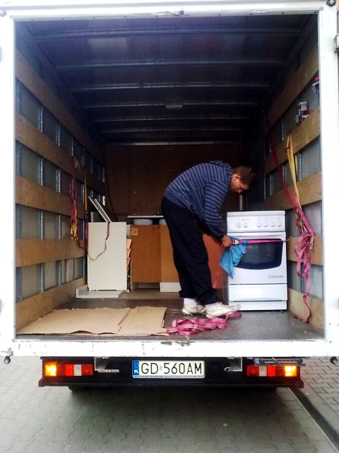 Wnętrze samochodu dostawczego: mężczyzna czyści kuchenkę przed transportem, widoczne kartony na podłodze i pasy zabezpieczające ładunek.