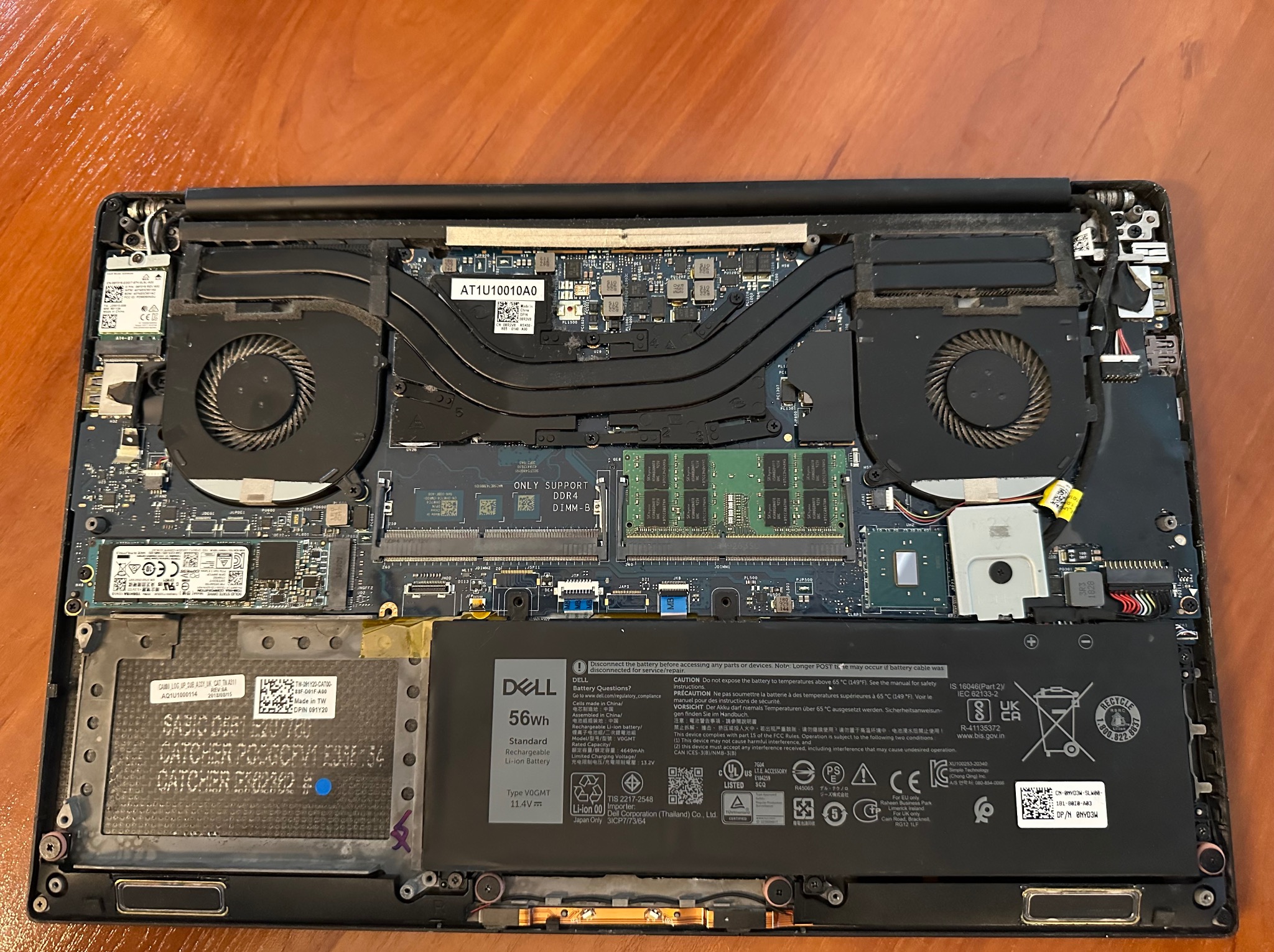 Otwarty laptop Dell z widocznymi komponentami wewnętrznymi: wentylatory, bateria, płyta główna, pamięć RAM. Widoczne oznaczenia i etykiety na podzespołach.