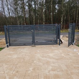 KL-Bruk - Nowoczesna, grafitowa brama przesuwna z poziomymi panelami, zamontowana na posesji z jasną kostką brukową. Widoczne drzewa w tle i fragment montażu.