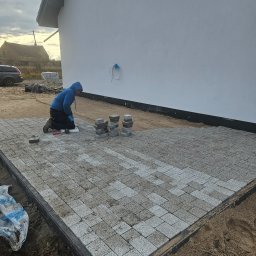KL-Bruk - Pracownik w niebieskiej bluzie klęczy układając szarą kostkę brukową na przygotowanym podłożu. Częściowo ułożona nawierzchnia, stosy kostki obok. Tło: biały budynek, auto.