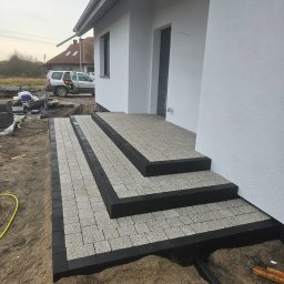 KL-Bruk - Schody wejściowe do domu wyłożone jasnoszarą kostką brukową z czarnym obramowaniem. Widoczne przygotowane podłoże i fragment domu z białym tynkiem.
