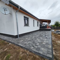 KL-Bruk - Nowoczesny taras z szarej kostki brukowej przy białym domu z ciemną podmurówką. Widoczny fragment ogrodu i pracujący robotnicy w tle. Ujęcie z niskiej perspektywy.