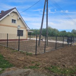 KL-Bruk - Nowoczesne ogrodzenie panelowe z podmurówką betonową, otaczające posesję z domem jednorodzinnym. Widoczne słupy i siatka w kolorze grafitowym, świeżo wyrównana ziemia wewnątrz ogrodzenia.