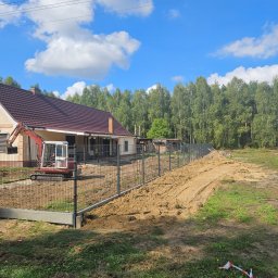 KL-Bruk - Nowoczesne ogrodzenie panelowe z podmurówką na nierównym terenie, z widocznym mini-koparką i domem w tle, w słoneczny dzień. Widoczne ślady prac ziemnych.