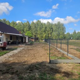 KL-Bruk - Montaż ogrodzenia panelowego na betonowej podmurówce, widok na posesję z domem i przygotowanym terenem pod trawnik. Słoneczny dzień, błękitne niebo.