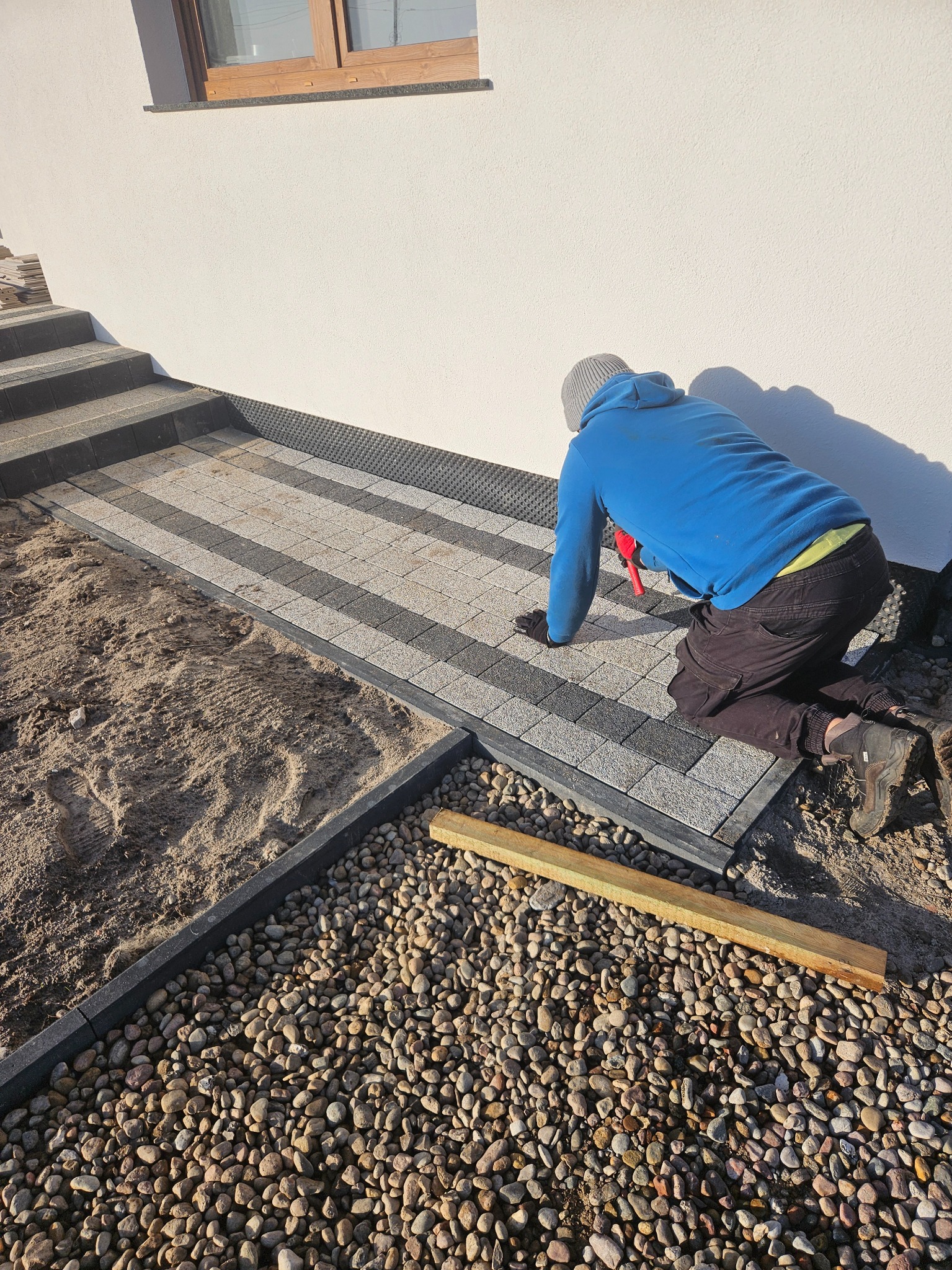 Pracownik w niebieskiej bluzie klęczy układając dwukolorową kostkę brukową przy białej ścianie domu. Widoczne kamienie ozdobne i schody.