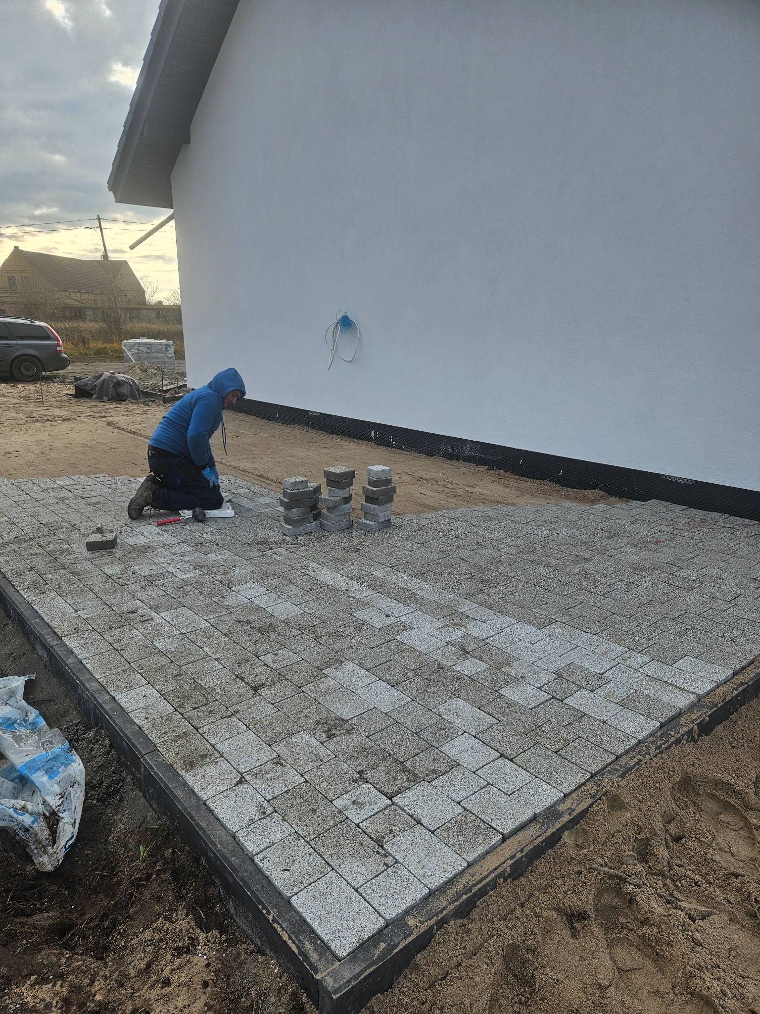 Pracownik w niebieskiej bluzie klęczy układając szarą kostkę brukową na przygotowanym podłożu. Częściowo ułożona nawierzchnia, stosy kostki obok. Tło: biały budynek, auto.