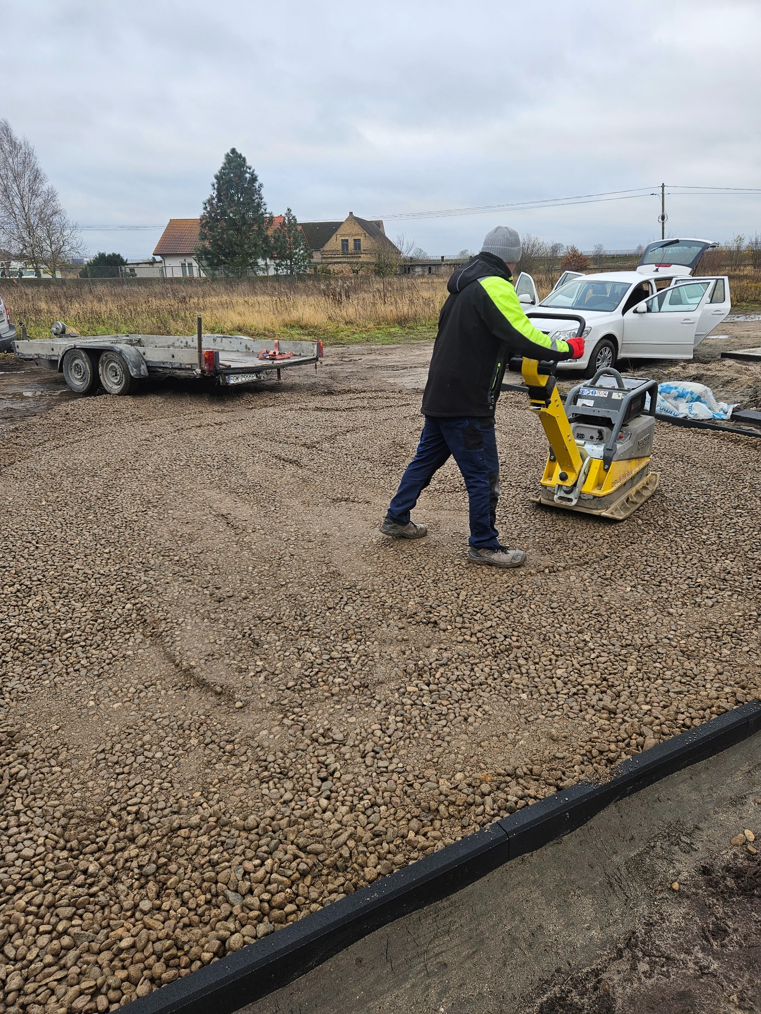 Pracownik w odblaskowej kurtce zagęszcza podłoże pod kostkę brukową za pomocą zagęszczarki. W tle przyczepa i samochód osobowy z otwartymi drzwiami.