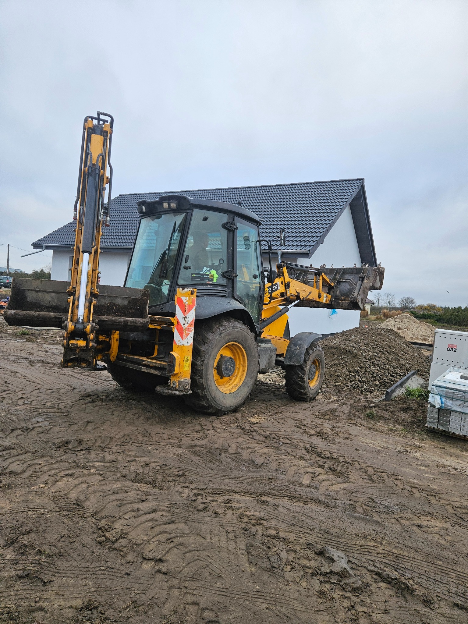 Żółta koparko-ładowarka na placu budowy z nowym domem w tle. Widoczne ślady opon w błocie i materiały budowlane. Praca w toku, przygotowanie terenu pod inwestycję.