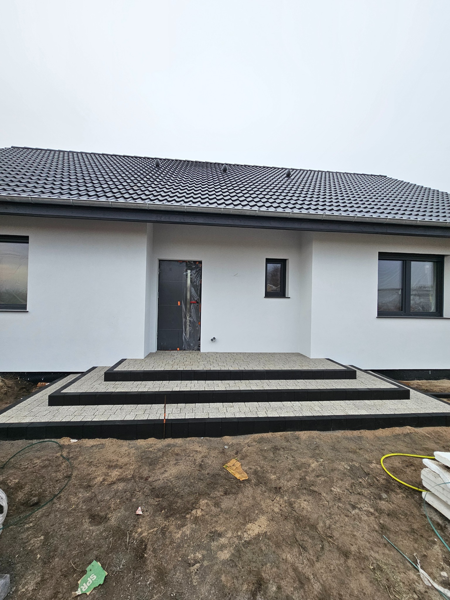 Nowoczesny dom z jasną elewacją i schodami wejściowymi wyłożonymi kostką brukową w odcieniach szarości i czerni. Minimalistyczny design, surowy krajobraz.