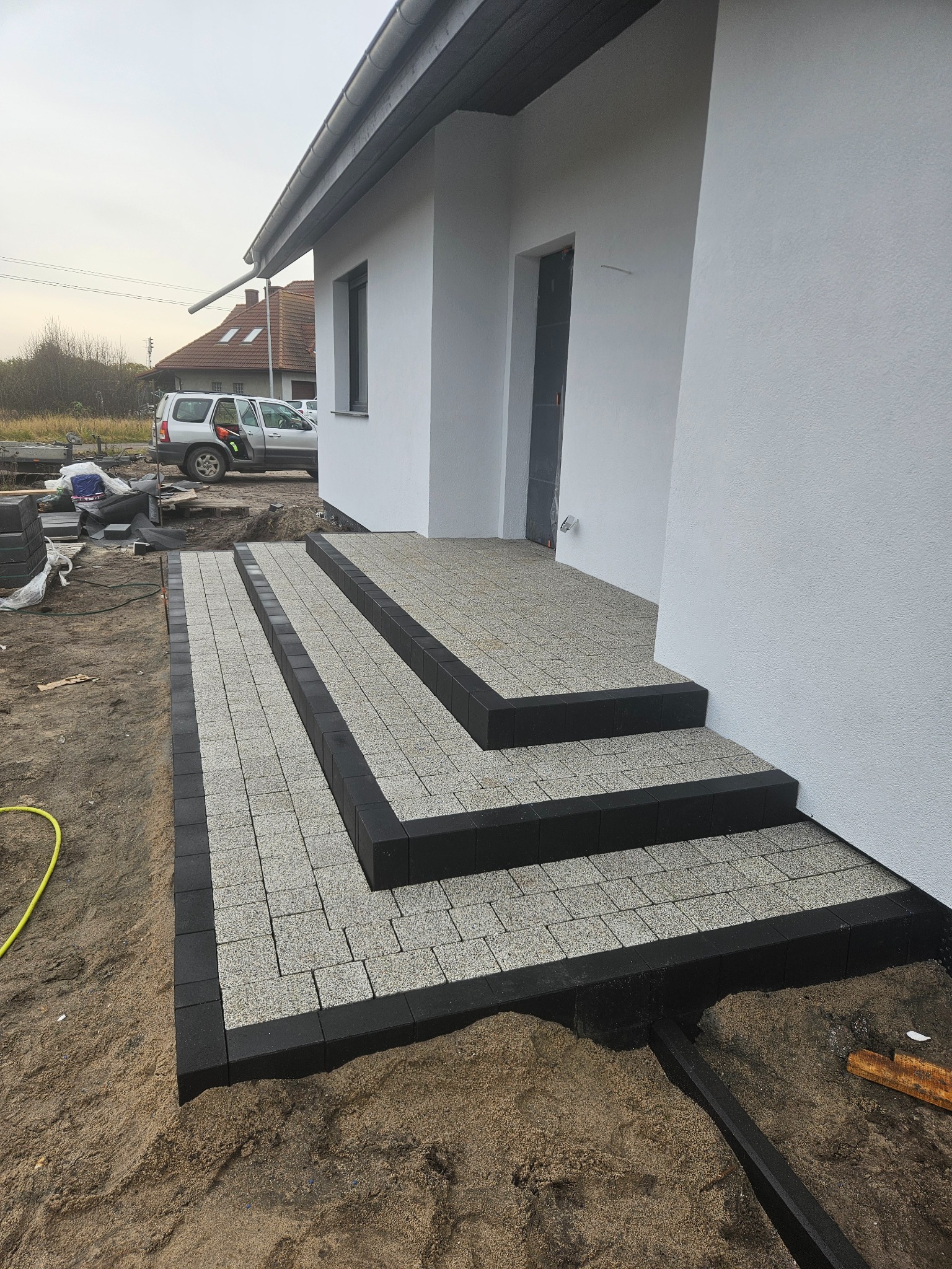Schody wejściowe do domu wyłożone jasnoszarą kostką brukową z czarnym obramowaniem. Widoczne przygotowane podłoże i fragment domu z białym tynkiem.