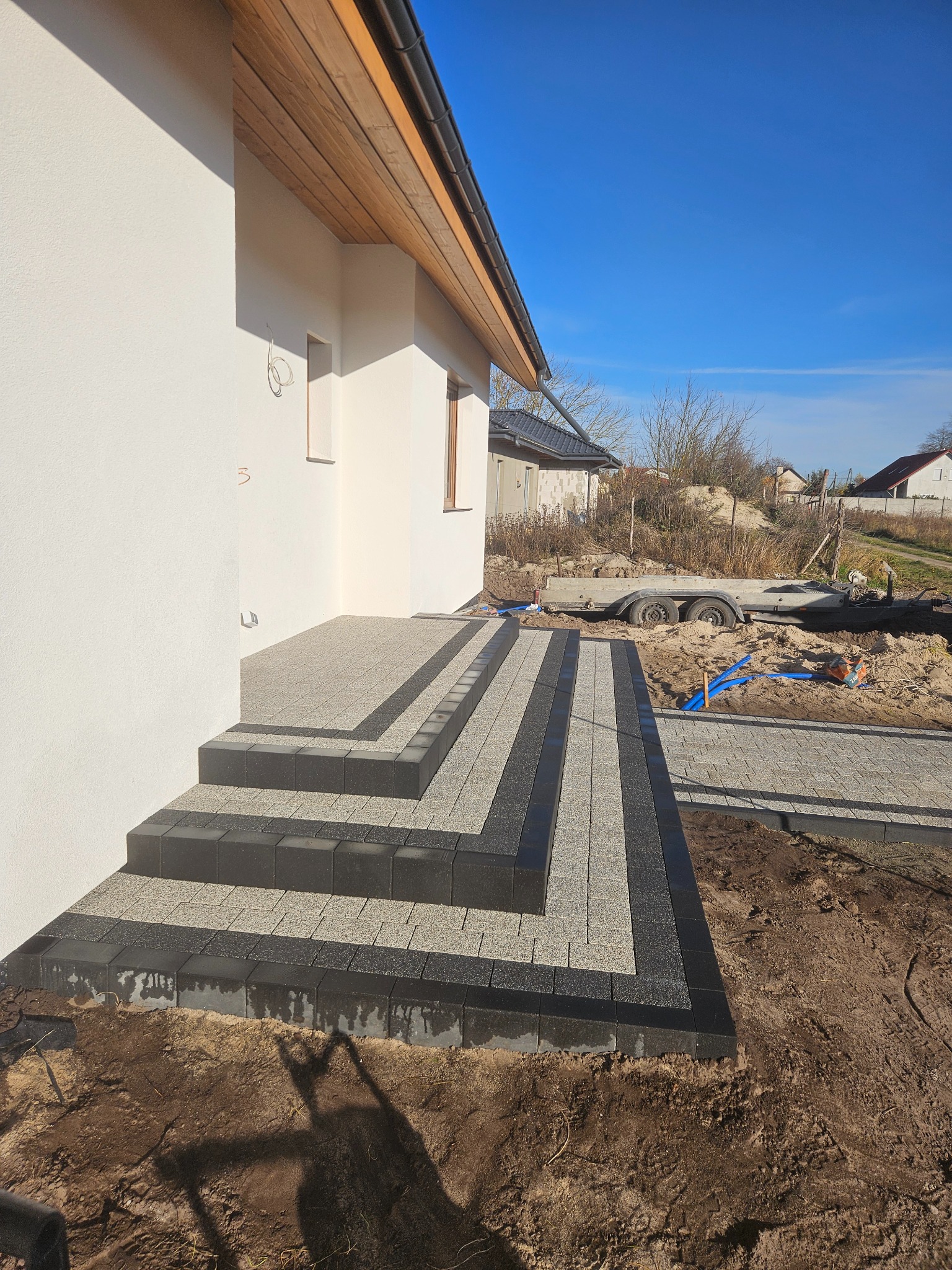 Nowoczesne schody wejściowe z kostki brukowej w odcieniach szarości, prowadzące do białego budynku z drewnianym podbitką. Widoczny fragment posesji i błękitne niebo.