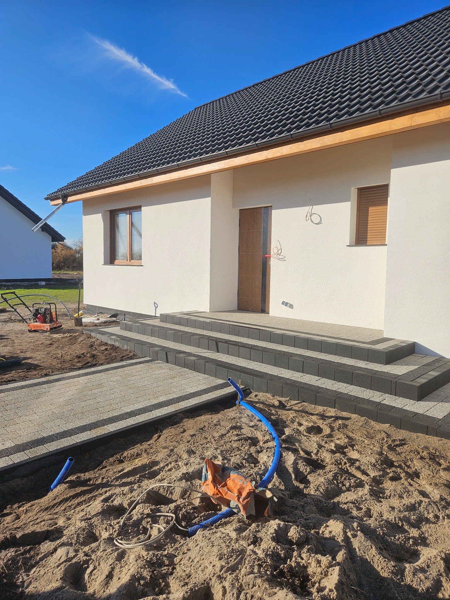 Nowoczesny dom z wybrukowanym wejściem i schodami z ciemnej kostki. Częściowo ułożona jasna kostka brukowa. Widoczne rury instalacyjne i zagęszczarka.