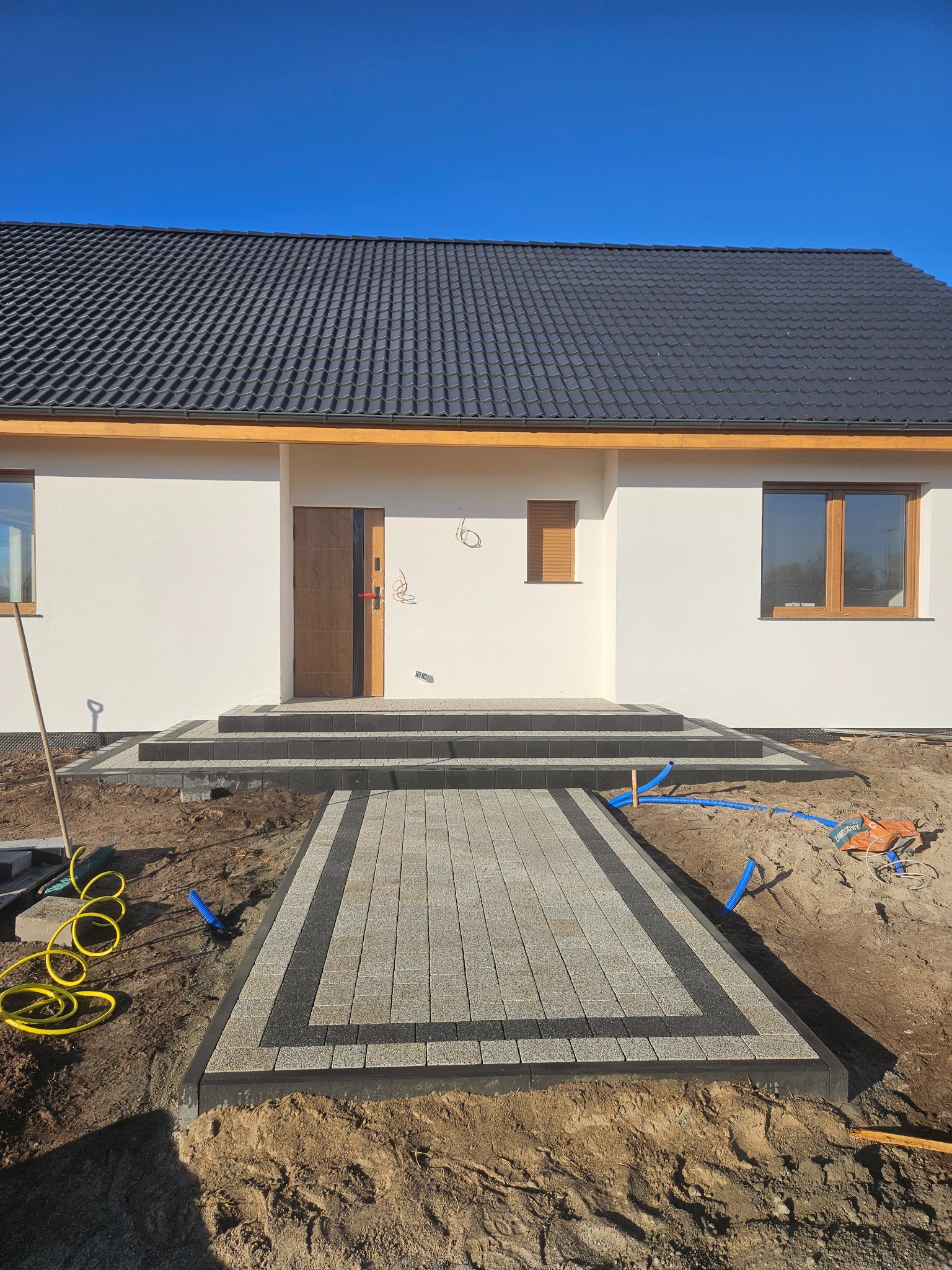 Nowoczesny dom z ułożoną kostką brukową w odcieniach szarości i czerni, prowadzącą do wejścia. Widoczne schody z kostki i fragment ogrodu w trakcie prac wykończeniowych.