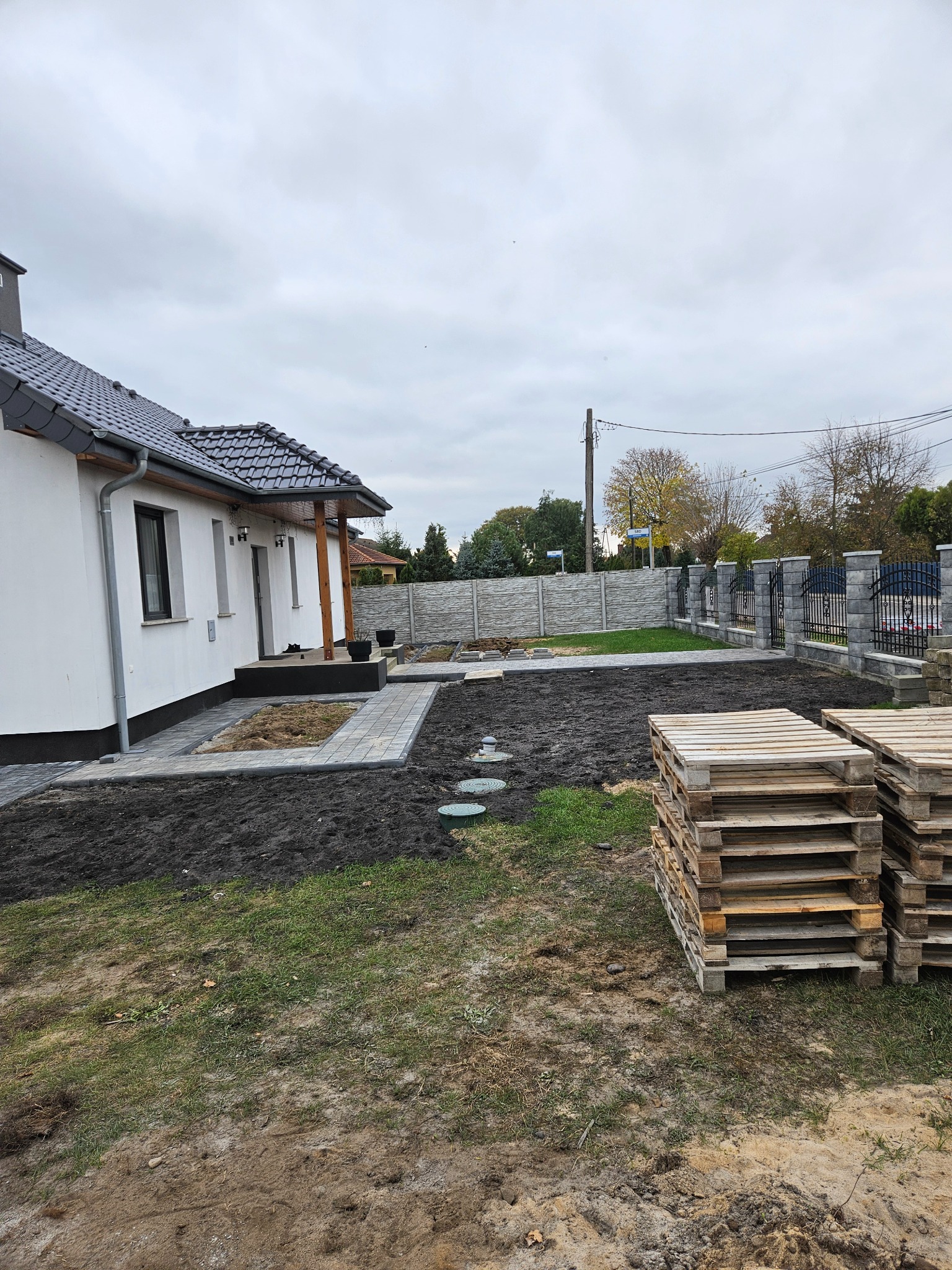 Ułożona kostka brukowa wokół domu i rabatki, widoczny fragment ogrodzenia z metalowymi przęsłami. Na pierwszym planie palety. Prace brukarskie w toku.