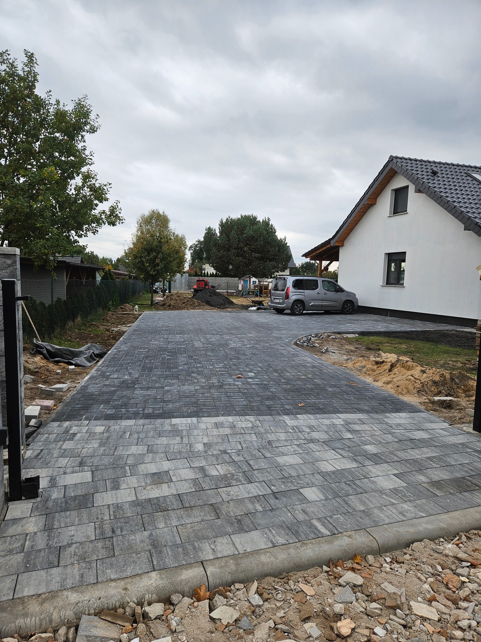 Ułożona, szara kostka brukowa na podjeździe do domu jednorodzinnego. Widoczny samochód i fragment ogrodzenia. Na obrzeżach kostki gruz i ziemia.