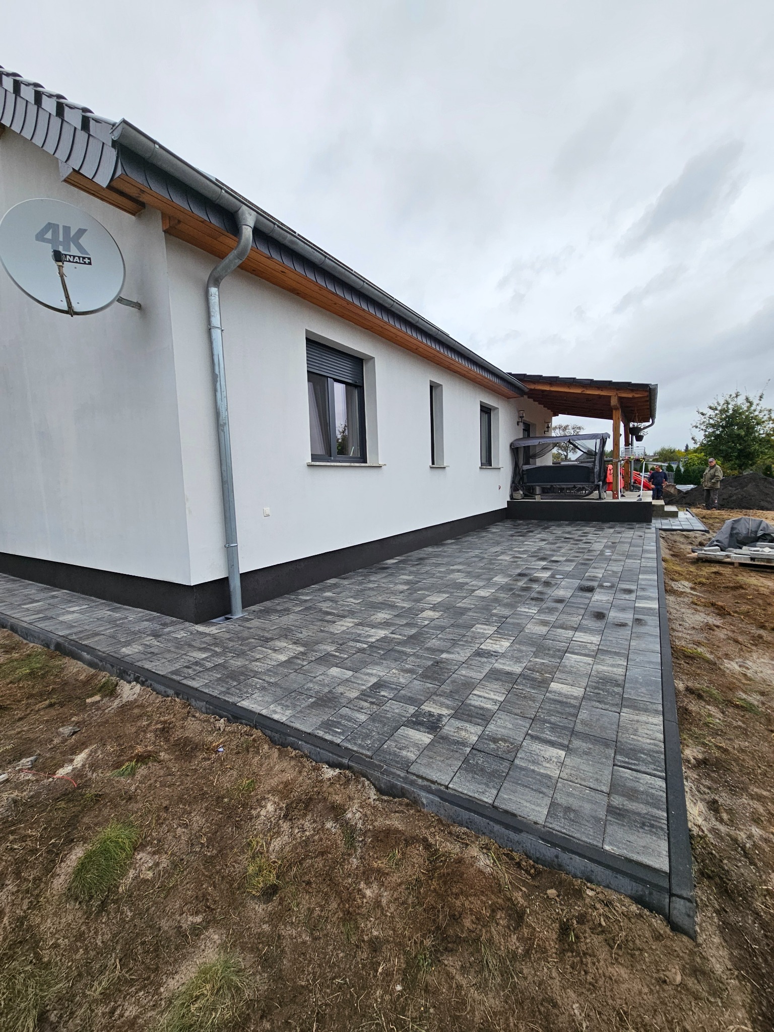 Nowoczesny taras z szarej kostki brukowej przy białym domu z ciemną podmurówką. Widoczny fragment ogrodu i pracujący robotnicy w tle. Ujęcie z niskiej perspektywy.