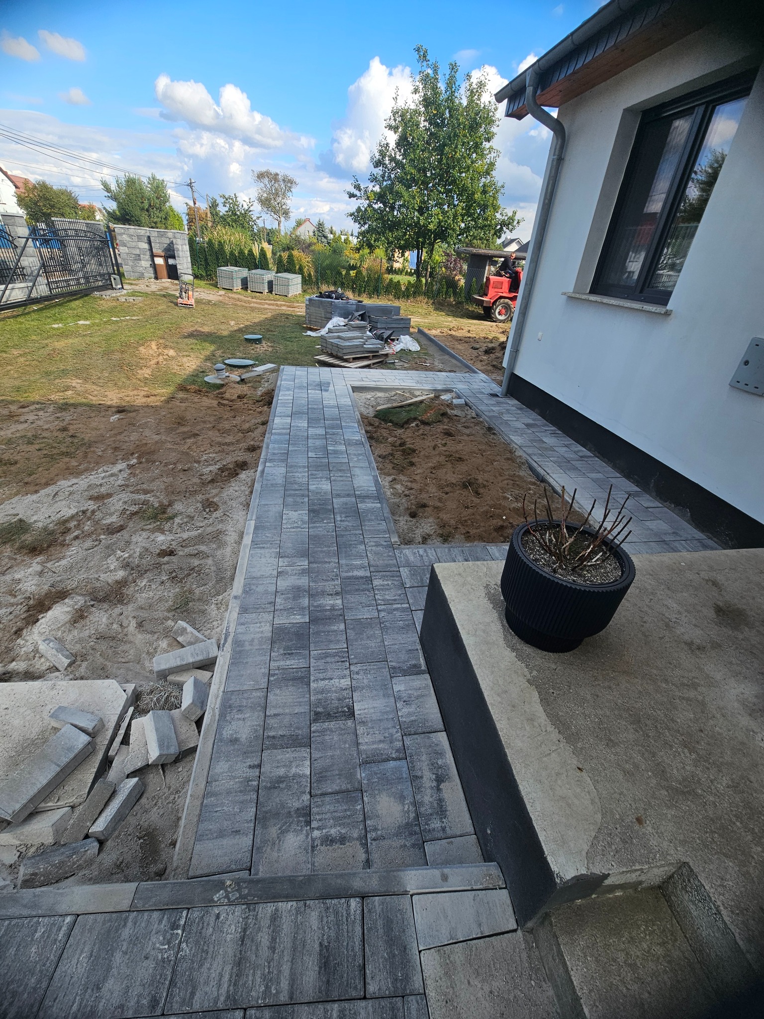 Fragment układania szarej kostki brukowej na posesji. Widoczne przygotowane podłoże, palety z kostką i budynek w tle. Częściowo ułożony chodnik z kostki.