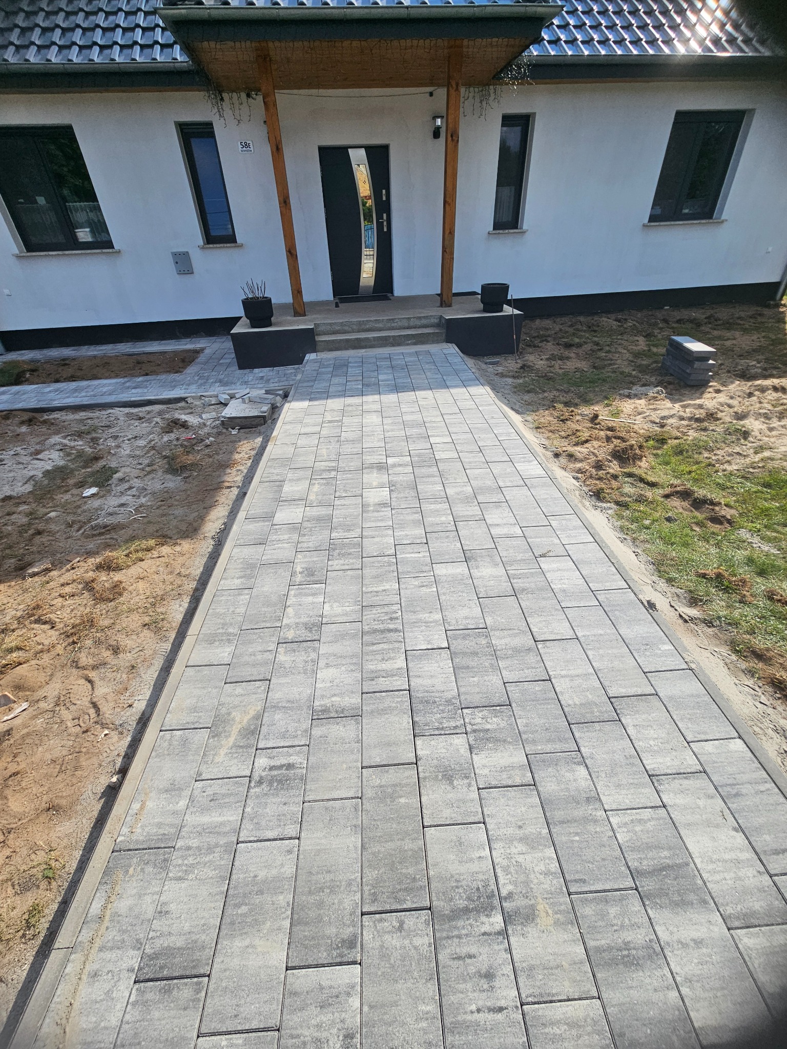 Ułożona ścieżka z szarej kostki brukowej prowadząca do domu z białymi ścianami i ciemnymi oknami. Widoczne detale wykończenia i fragmenty niezabrukowanego terenu.
